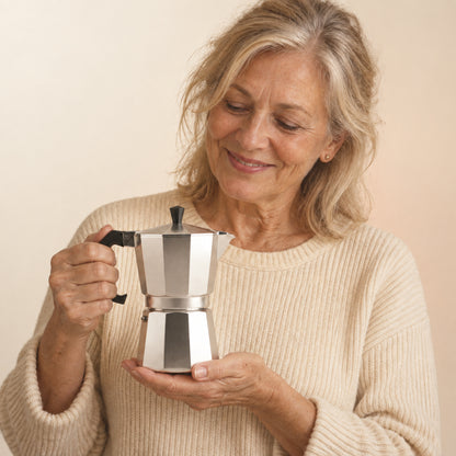 An older blonde woman in a cream knit sweater holding the octagonal aluminum La Moka pot against a soft cream, sand and peach studio gradient.